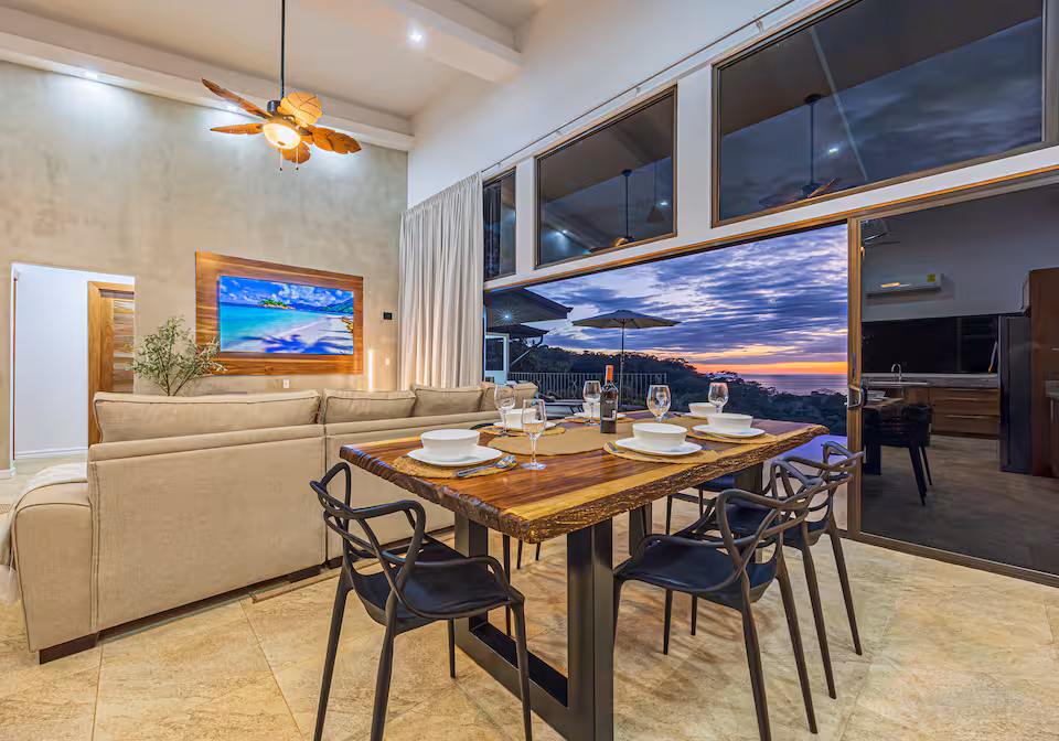 Indoor dining table set for dinner beside floor-to-ceiling windows at dusk in KsaLinda villa, Costa Rica