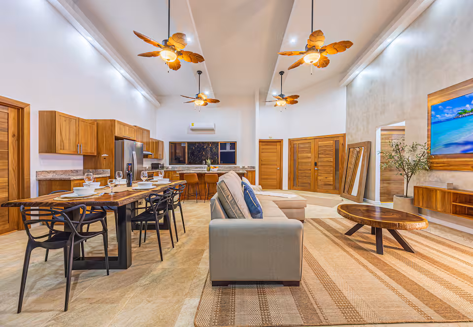 Open-plan great room at KsaLinda with vaulted ceiling, palm-blade fans, dining table, sofa and kitchen beyond